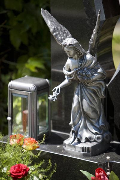 Silberne Engel-Skulptur mit Blumenstrauß neben Glas-Laterne auf einem dunklen Granit-Grabstein